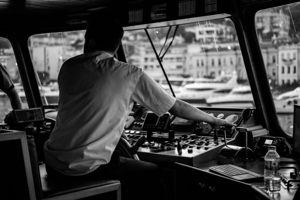 An atmospheric black and white image of a captain navigating a vessel in a busy harbor environment.